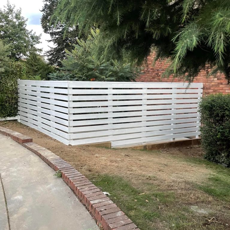 A white slatted fence surrounding a small grassy area near a brick wall.